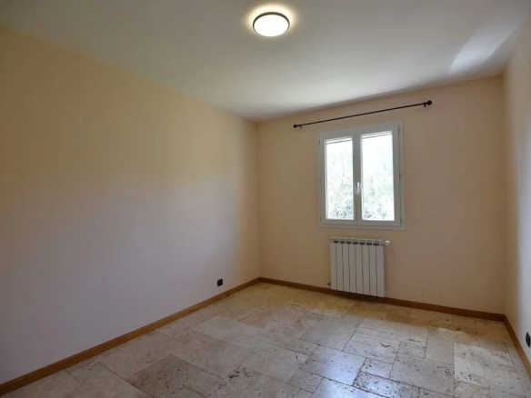 Empty beige room with tiled floor, a window on the far wall, and a wall-mounted radiator beneath it; a curtain rod above the window; ceiling light on.