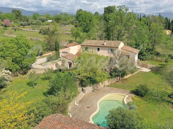 A hillside villa with a tiled roof and beige walls, surrounded by trees and stone walls, with a curved swimming pool in the foreground.