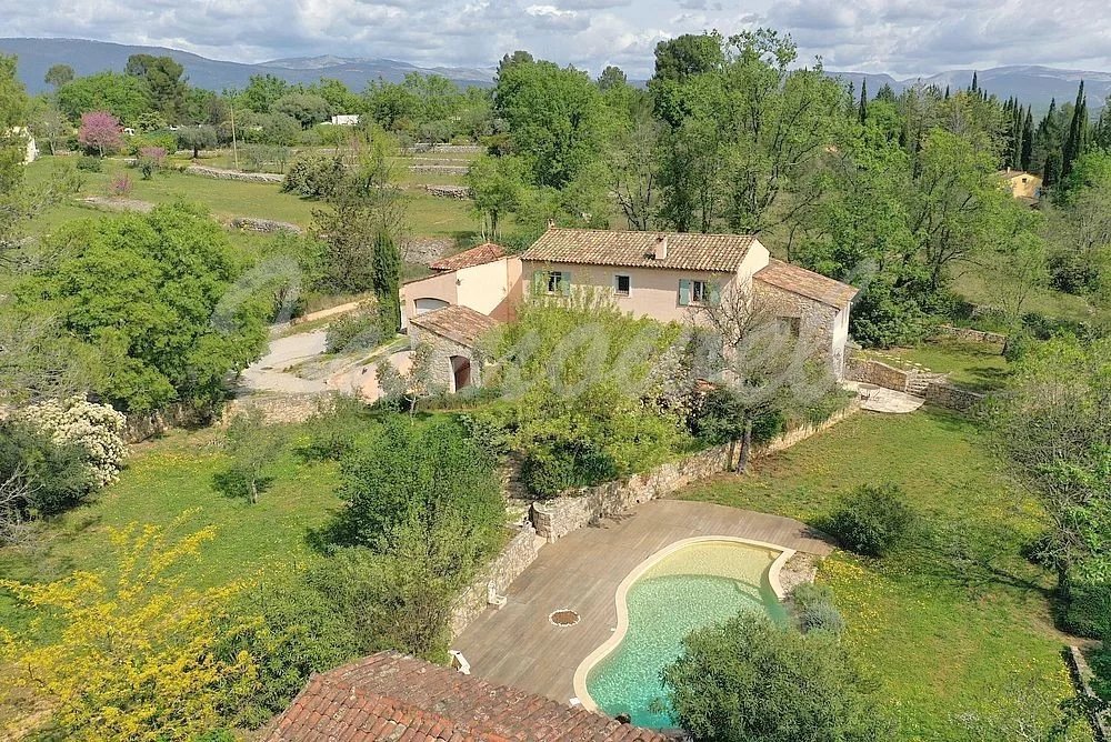 A hillside villa with a tiled roof and beige walls, surrounded by trees and stone walls, with a curved swimming pool in the foreground.