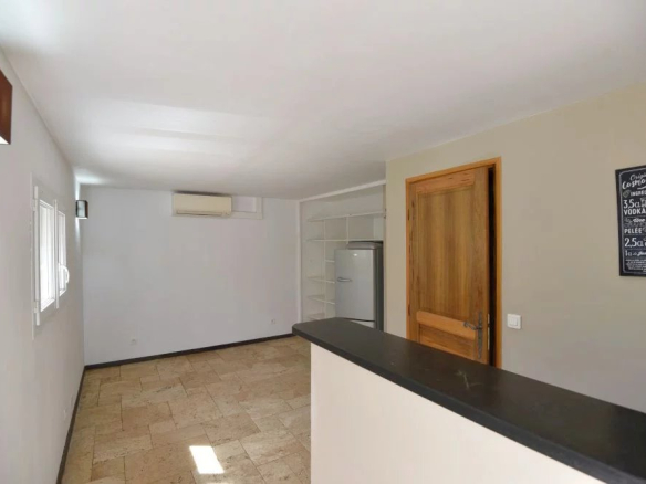 Interior of a small kitchen with a dark breakfast-bar counter in the foreground, beige tiled floor, white walls, a fridge and open shelves against the back wall, a wooden door on the right, and a chalkboard sign on the wall.