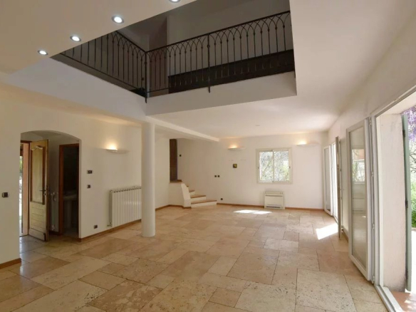 Bright, empty living room with beige tile floor, white walls, large sliding glass doors to a garden, and a wrought-iron railing on a mezzanine above.
