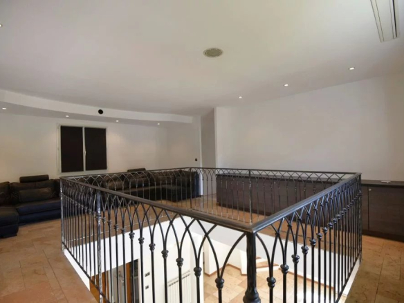 Open living area with a black wrought-iron railing around a stairwell, tan tile floor, and dark leather sectional against white walls.