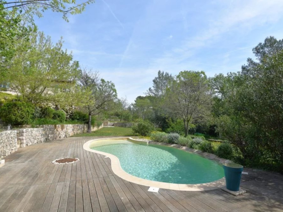 Kidney-shaped pool with turquoise water on a wooden deck, surrounded by stone walls and green trees on a sunny day.