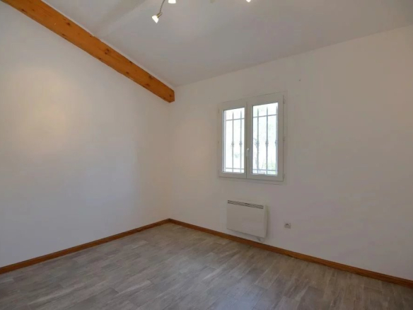 Empty white-walled room with a wooden ceiling beam, gray wood-look flooring, a window with bars, and a wall heater beneath it.