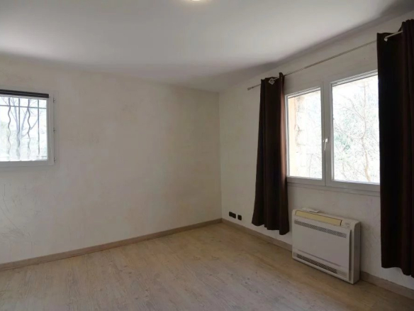 Empty rectangular living room with light wood flooring, white walls, and two windows with dark curtains; a wall heater beneath the right window.