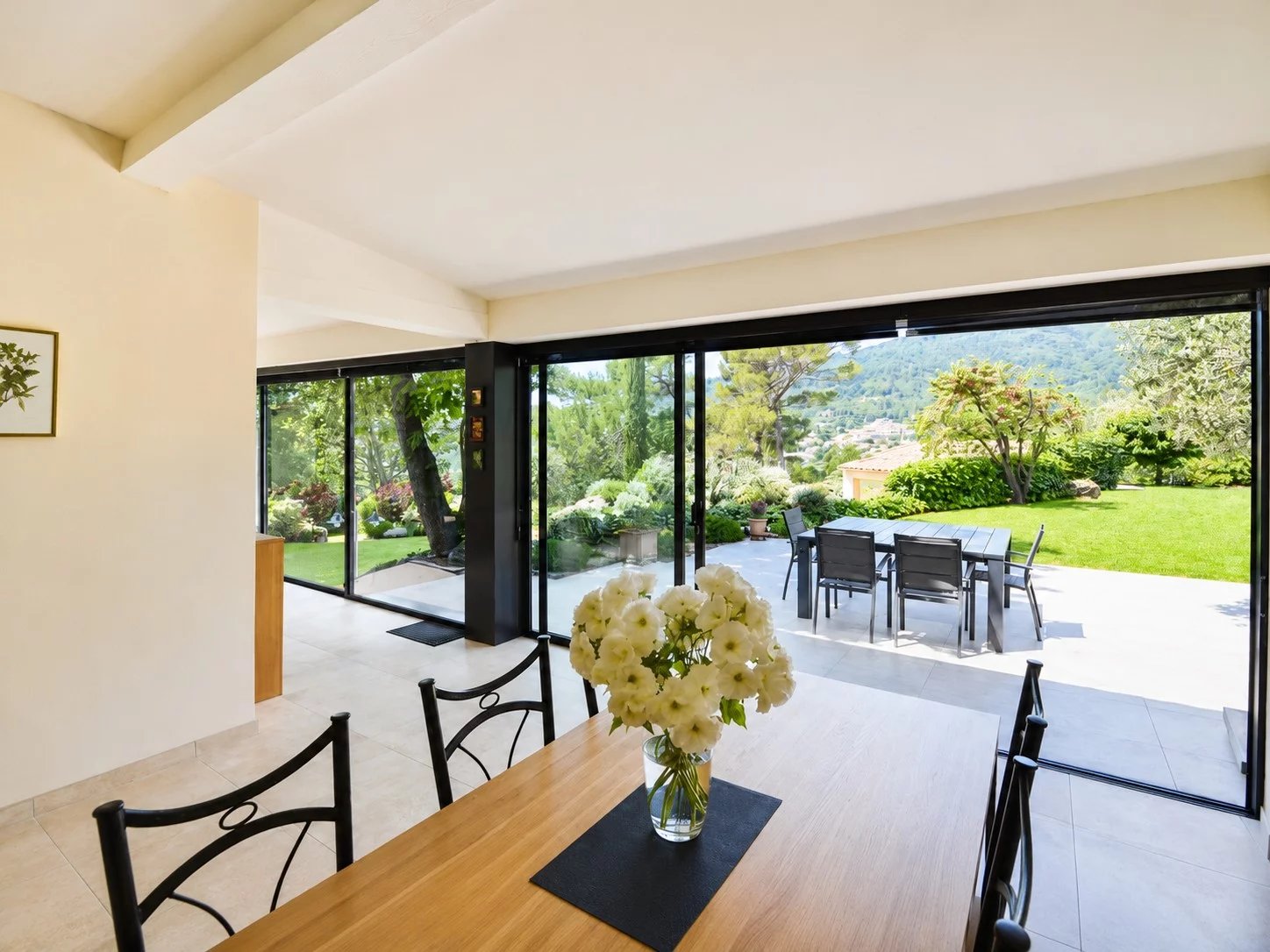 Intérieur d'une villa avec vue sur un jardin et des collines, table en bois et fleurs blanches.
