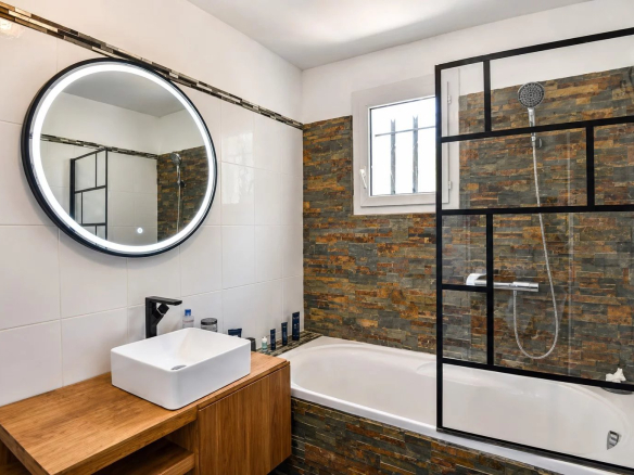 Salle de bain moderne avec baignoire et miroir rond, carrelage en pierre et meuble en bois.