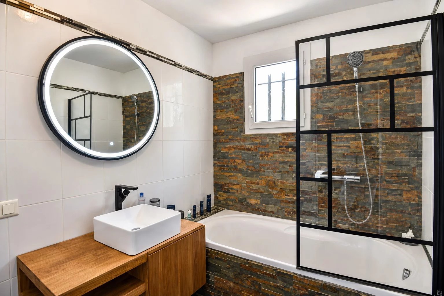 Salle de bain moderne avec baignoire et miroir rond, carrelage en pierre et meuble en bois.