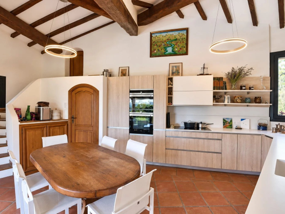 Modern kitchen with wooden table and white chairs, exposed beams and windows to the outside.
