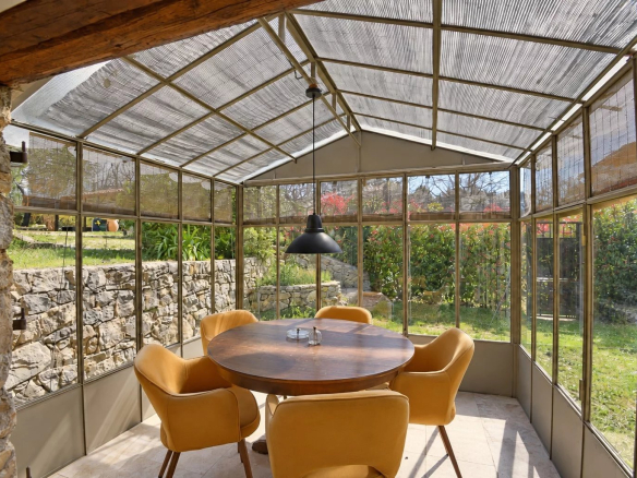 Véranda en verre avec table ronde et chaises, entourée de verdure.