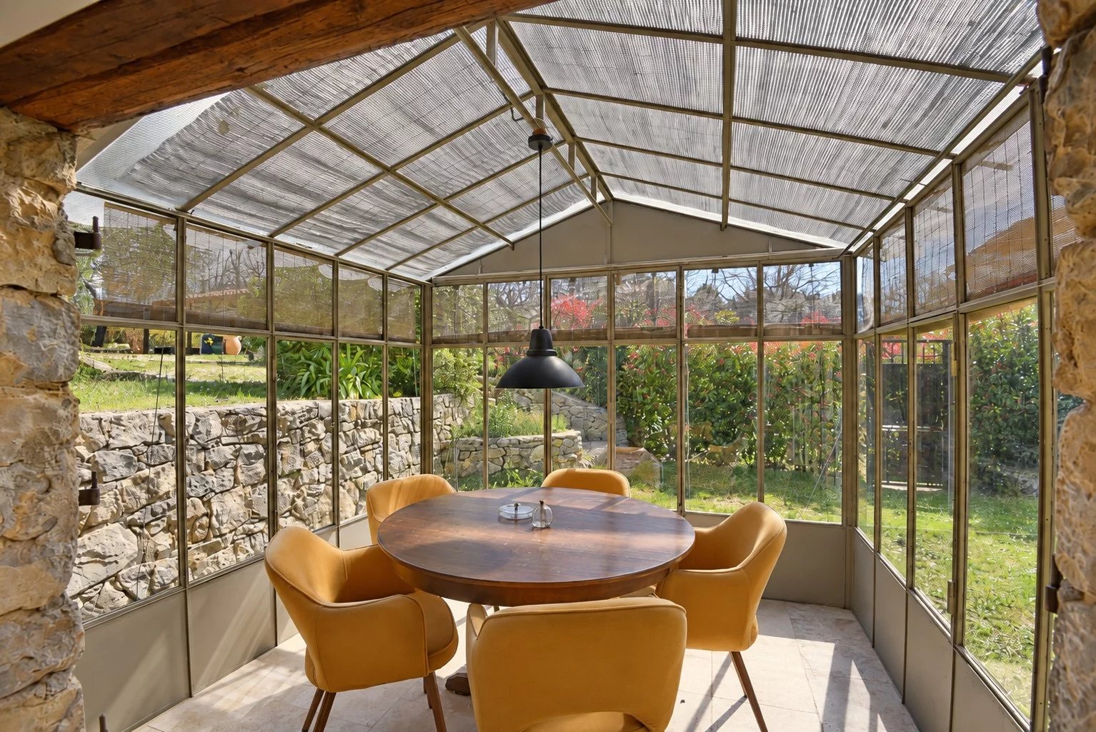 Véranda en verre avec table ronde et chaises, entourée de verdure.