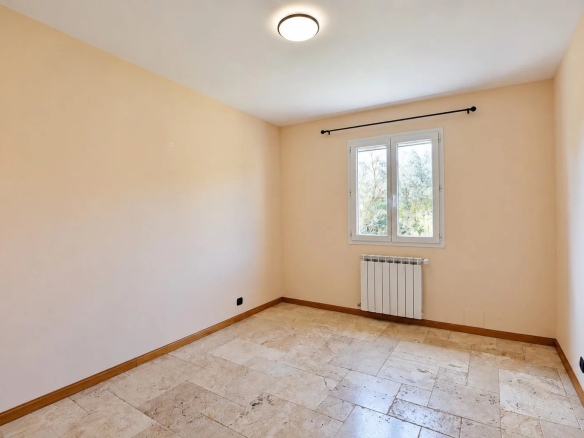 Chambre vide avec murs clairs et fenêtre, sol en pierre, radiateur visible.