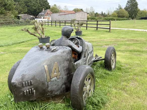 Statue d'un pilote dans une voiture de course ancienne dans un champ verdoyant près de Pont-l'Évêque.