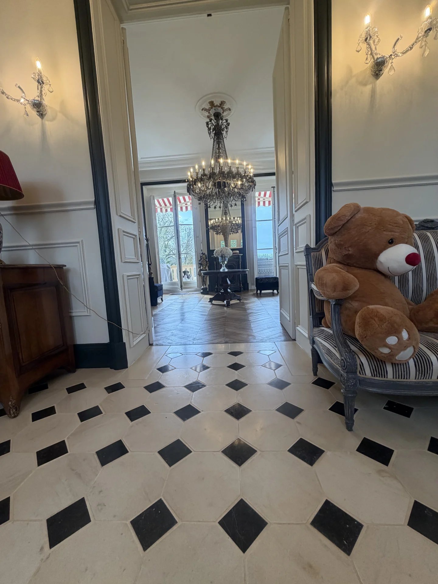 Intérieur d'un château avec un sol en carreaux noirs et blancs, un lustre et un ours en peluche sur une chaise.