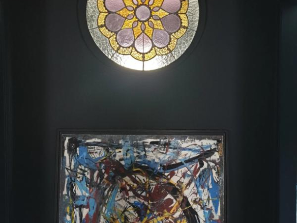 Circular stained-glass window with pink and yellow petal motif above a chaotic multicolored abstract painting leaning against a dark wall.