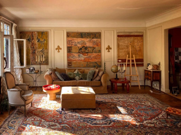 Sunlit living room with a tan sofa, patterned rug, and vintage chairs, plus wall art and an easel in the corner.