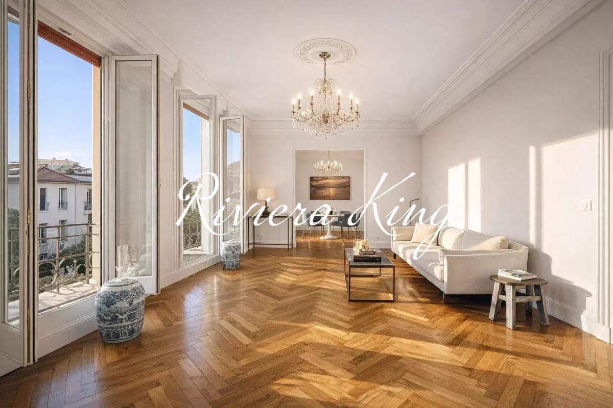 Bright living room with tall windows, herringbone wooden floor, and a crystal chandelier above a cream sofa set.