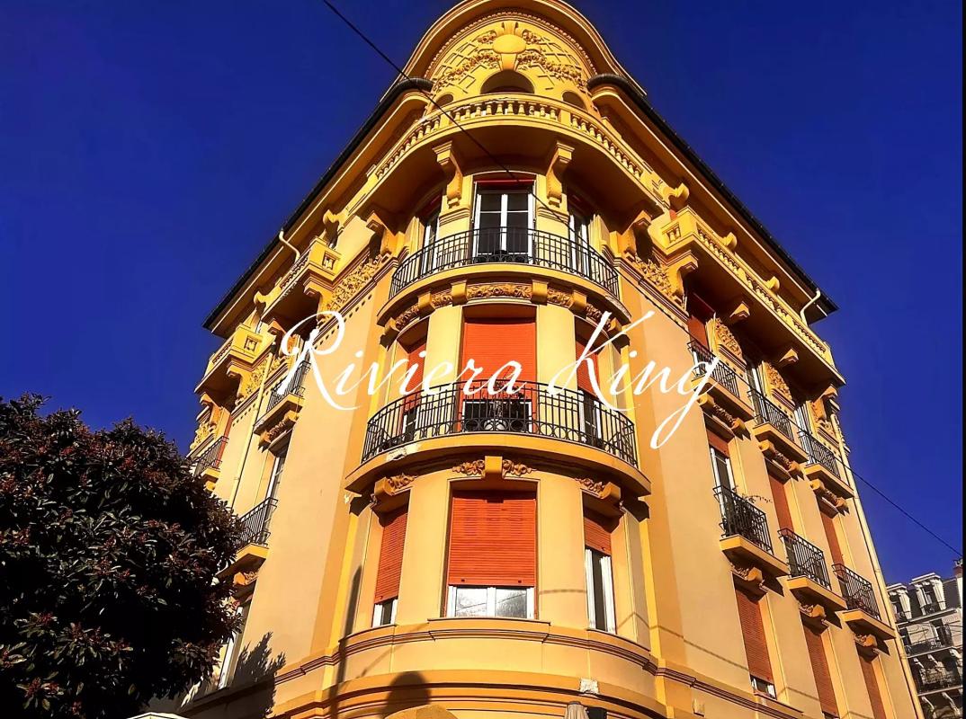 Yellow ornate corner apartment building with curved balconies, red shutters, and decorative details against a clear blue sky.