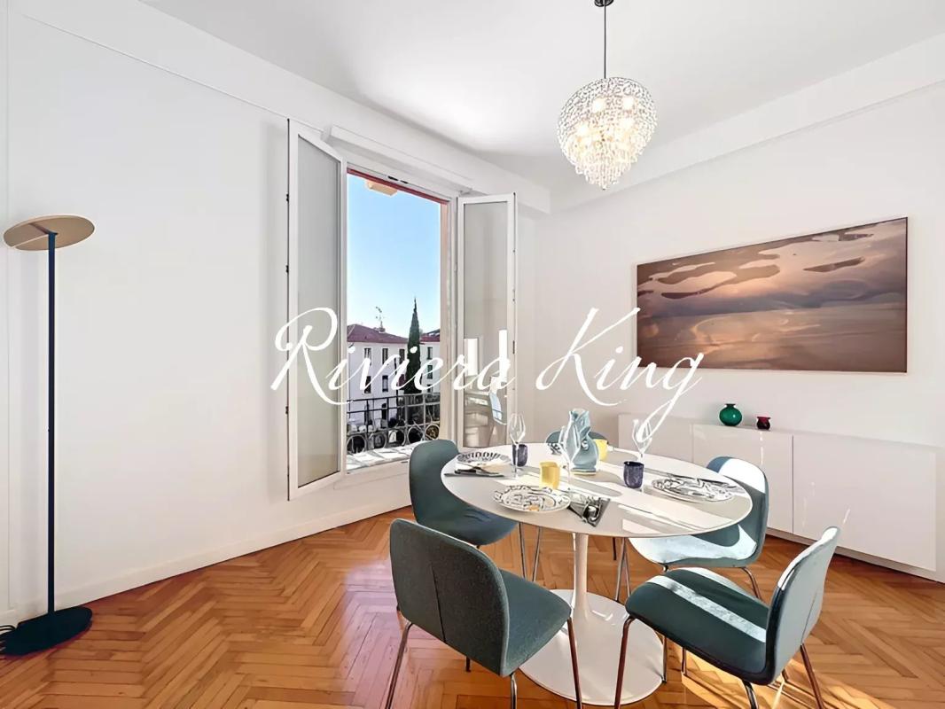 Bright dining room with a round white table set for four, teal chairs, open French doors to a balcony, and a crystal pendant light above.