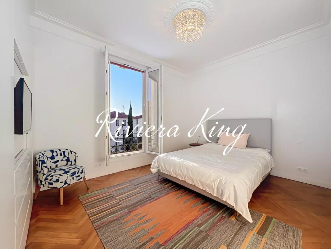 Bright white master bedroom with a large open window, chandelier, and a king bed with white bedding on a gray upholstered headboard, plus a patterned chair and colorful rug on hardwood floors.
