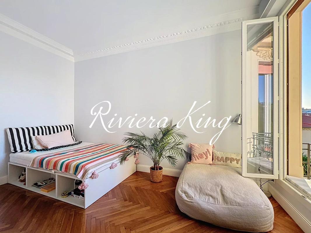 Bedroom with a white wall, bed with colorful striped bedding and built-in shelves underneath, a potted palm, and a large cushioned lounge by an open window leading to a balcony; overlay text reads 'Riviera King'.