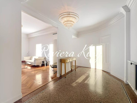 Bright foyer with a speckled terrazzo floor, white walls, and a large crystal chandelier; an opening leads to a sunlit living room with a white sofa and wooden floor, plus a small console table along the wall.