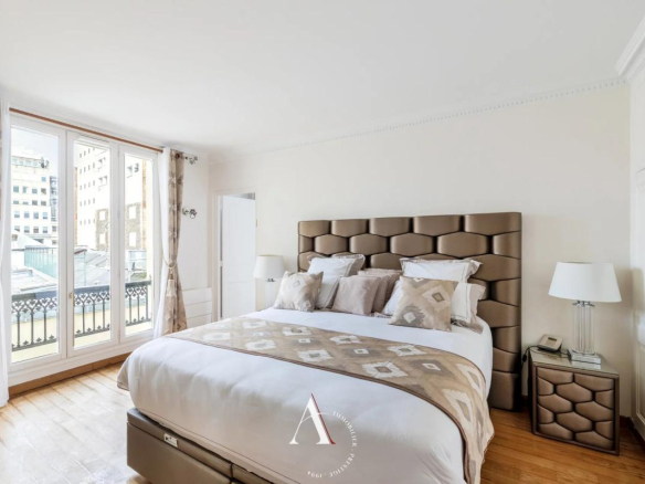 Bright hotel-style bedroom with a tall, tufted headboard, white bedding, and a beige patterned throw; large glass doors to a balcony on the left.