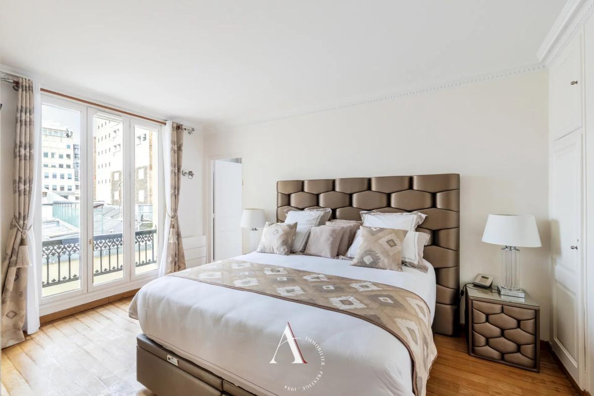 Bright hotel-style bedroom with a tall, tufted headboard, white bedding, and a beige patterned throw; large glass doors to a balcony on the left.