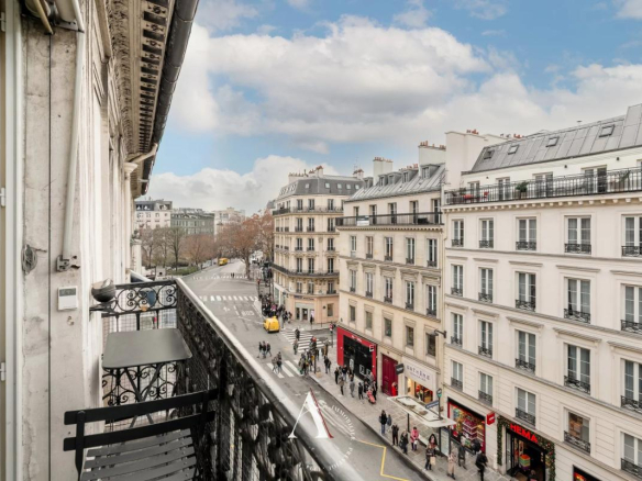 View from a balcony over a Parisian street lined with Haussmann buildings and a busy crosswalk below.