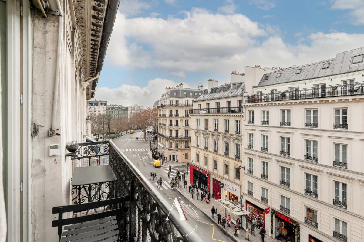 View from a balcony over a Parisian street lined with Haussmann buildings and a busy crosswalk below.