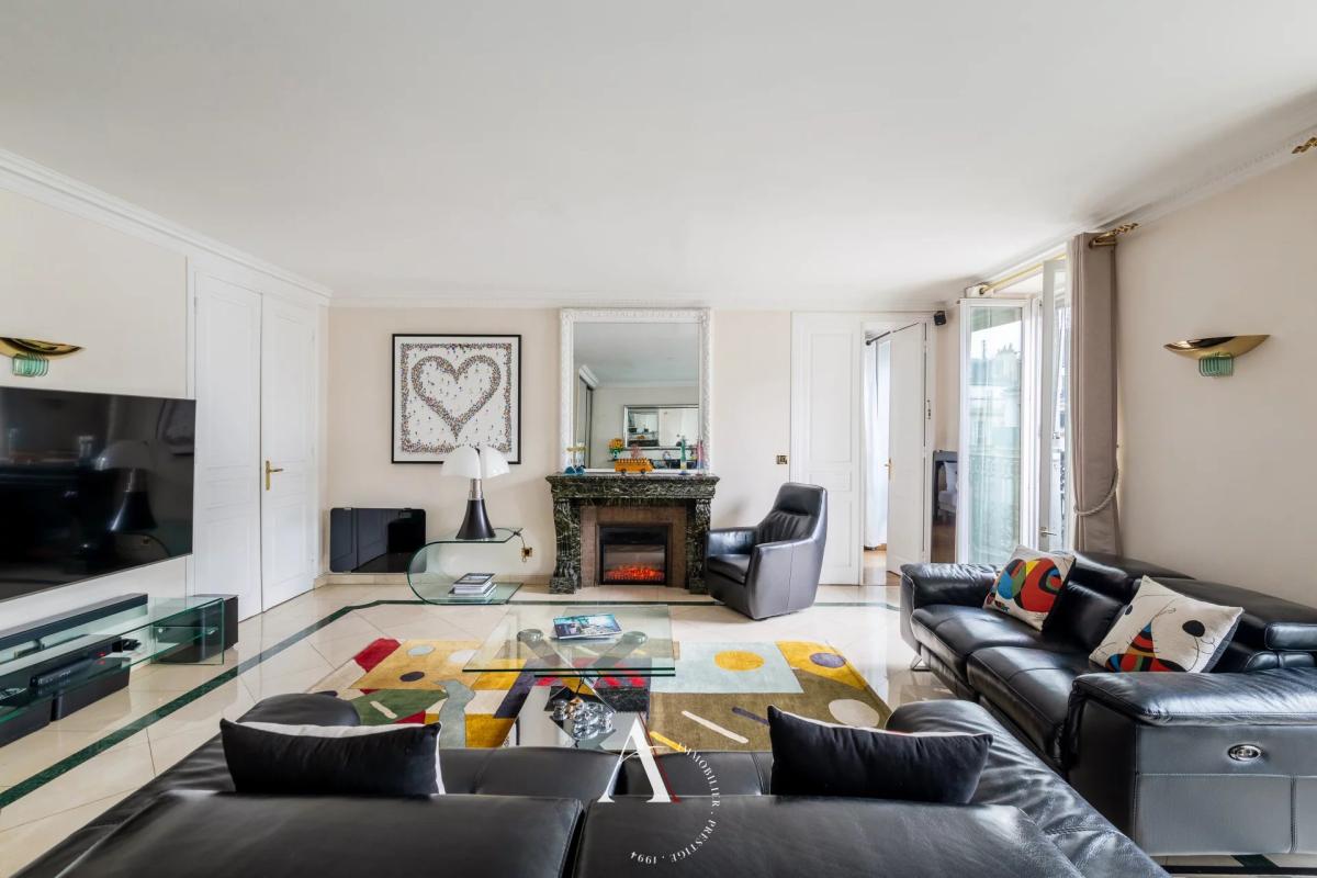 Bright living room with black leather sofas, glass coffee table, colorful rug, and a marble fireplace in a bright open plan space.