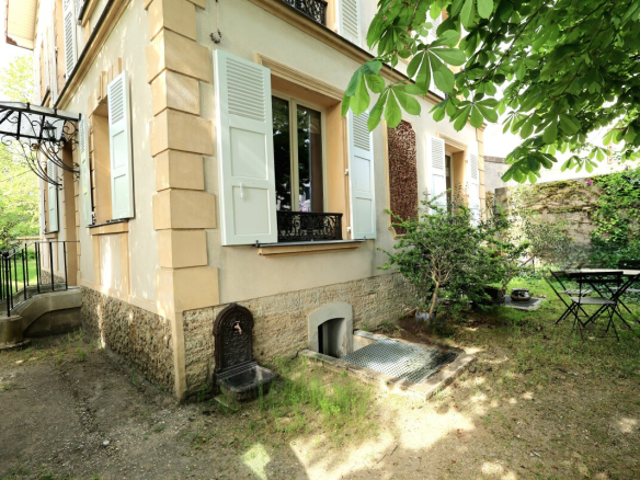 Beige house with pale blue shutters, a small garden, and a metal patio table and chairs under a leafy tree.