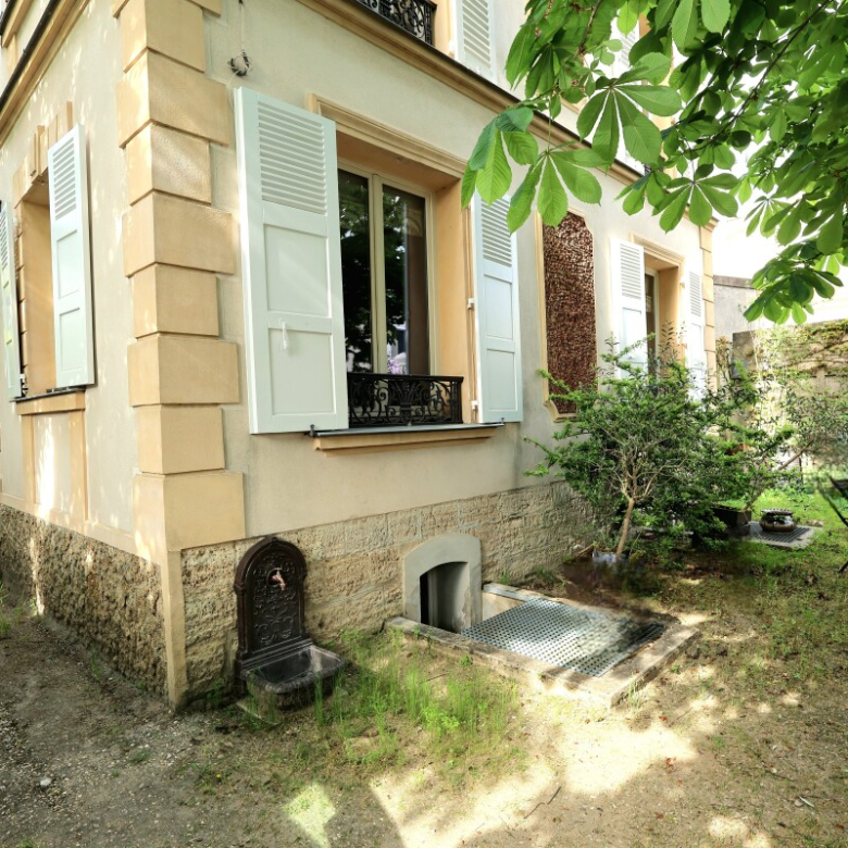 Beige house with pale blue shutters, a small garden, and a metal patio table and chairs under a leafy tree.