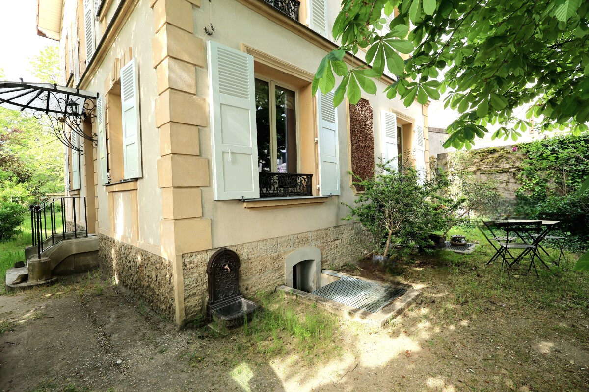 Beige house with pale blue shutters, a small garden, and a metal patio table and chairs under a leafy tree.