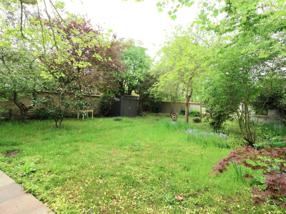 A green backyard with trees, a small shed along a fence, and a grassy lawn surrounded by flowering plants and shrubs.