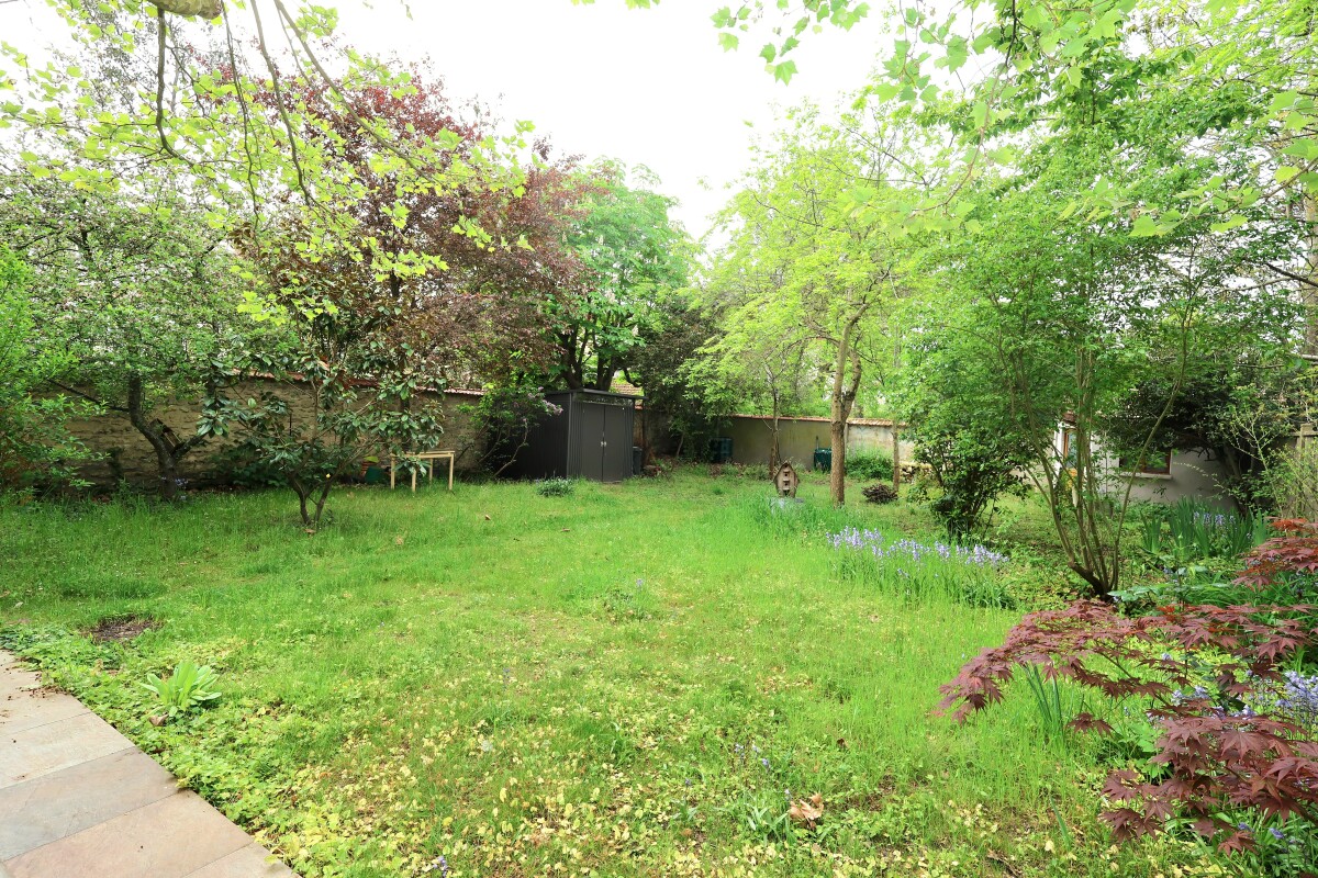 A green backyard with trees, a small shed along a fence, and a grassy lawn surrounded by flowering plants and shrubs.