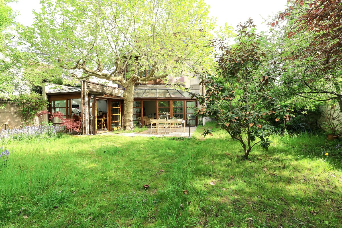 Garden with a lush lawn and a large tree in front of a glass-walled house; patio dining table and chairs outside.