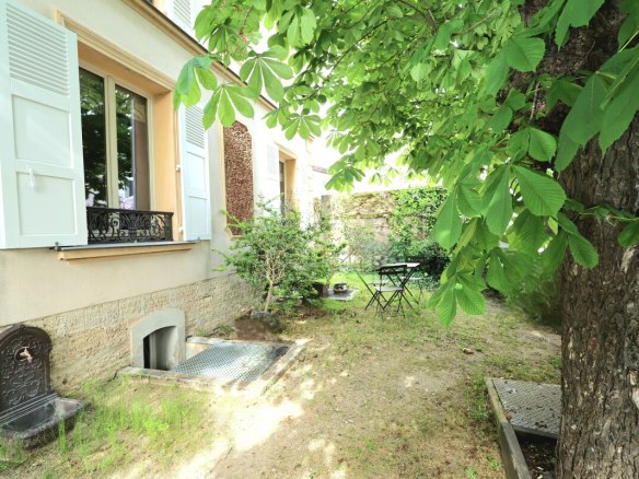 Cottage courtyard with pale green window shutters, a small door in the stone wall, and a leafy tree shading a lawn with a metal bistro table and chairs.