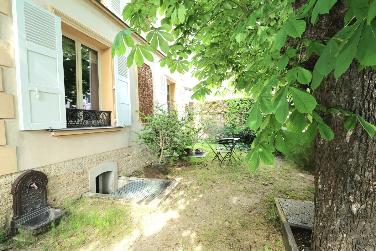 Cottage courtyard with pale green window shutters, a small door in the stone wall, and a leafy tree shading a lawn with a metal bistro table and chairs.