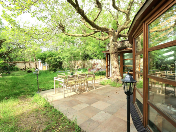 Patio with a metal dining table and chairs under a large tree, beside a glass-walled house in a green garden.
