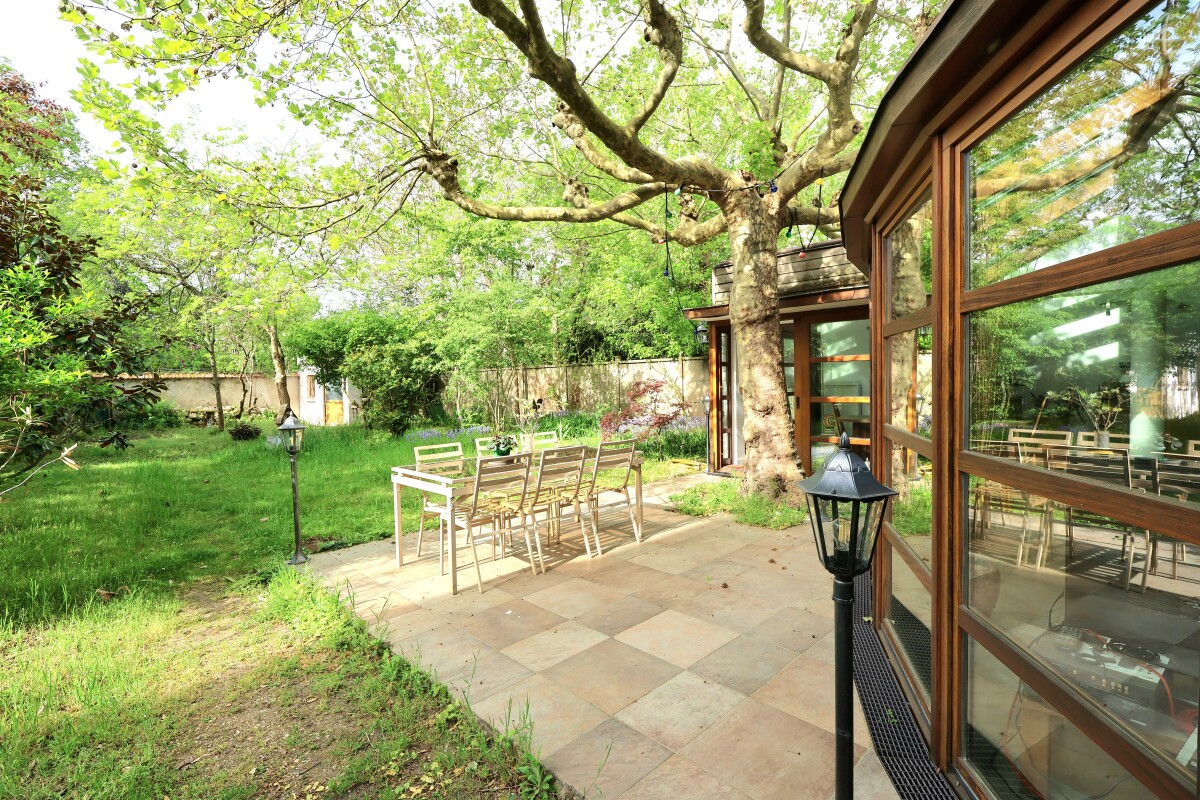 Patio with a metal dining table and chairs under a large tree, beside a glass-walled house in a green garden.