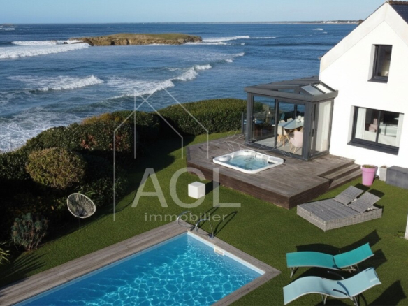 A coastal backyard with a rectangular blue pool, wooden deck, hot tub, and sun loungers beside a white house overlooking the sea.