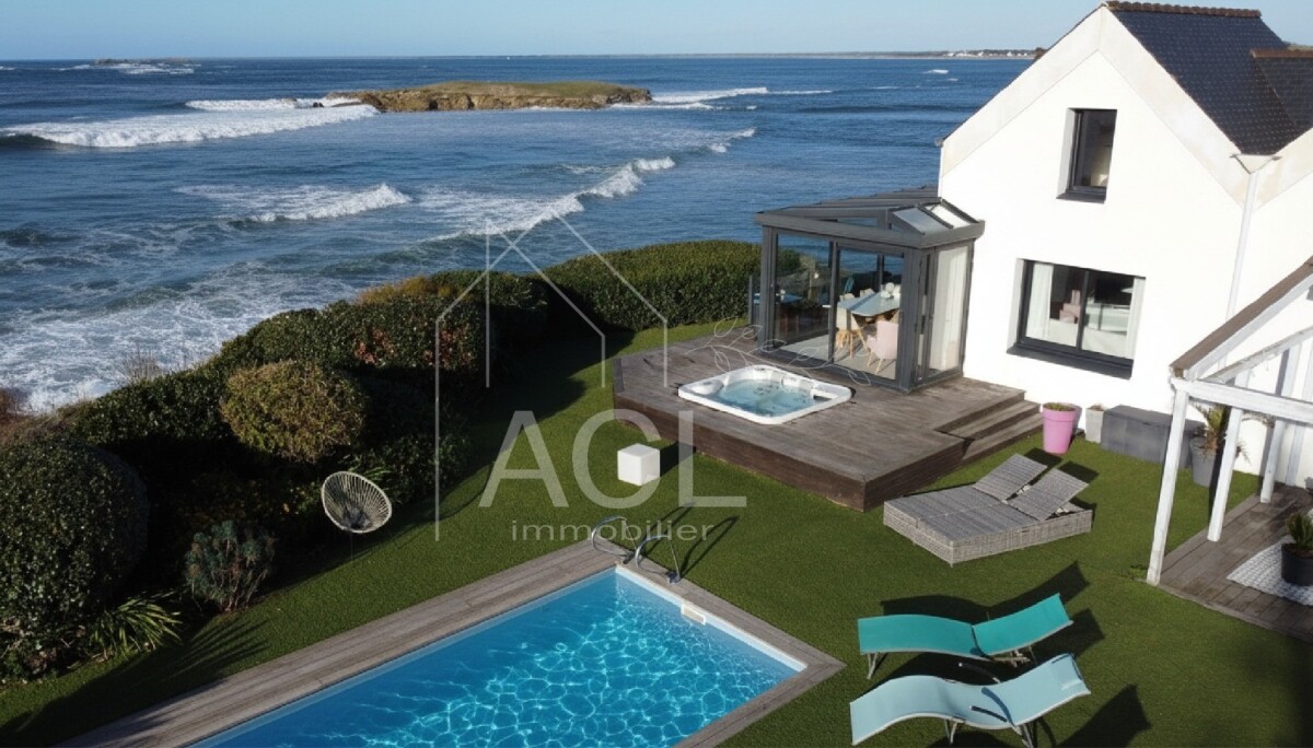 A coastal backyard with a rectangular blue pool, wooden deck, hot tub, and sun loungers beside a white house overlooking the sea.