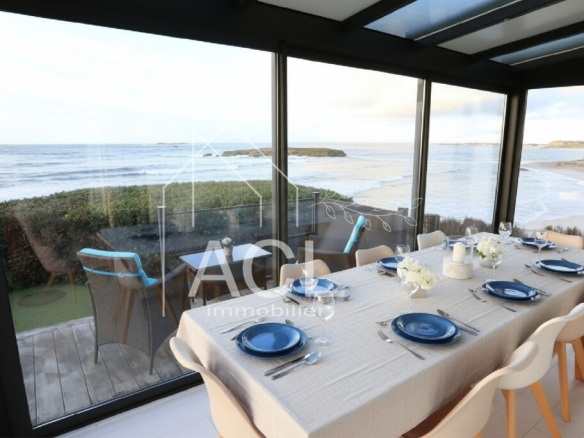 Dining table set for eight in a glass-walled seaside dining room with a beach and ocean view outside.