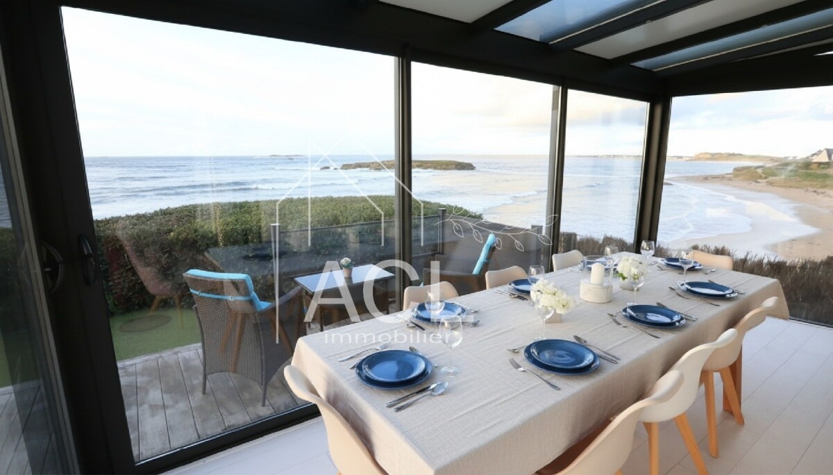 Dining table set for eight in a glass-walled seaside dining room with a beach and ocean view outside.