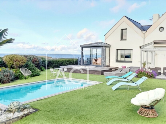White modern house with a glass conservatory, a rectangular pool, and a sunlit lawn with loungers and seating, sea view in the background.