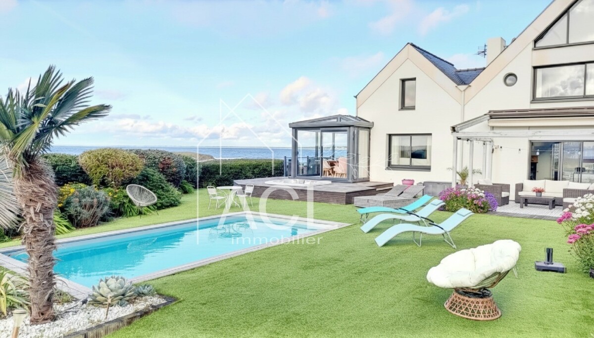 White modern house with a glass conservatory, a rectangular pool, and a sunlit lawn with loungers and seating, sea view in the background.