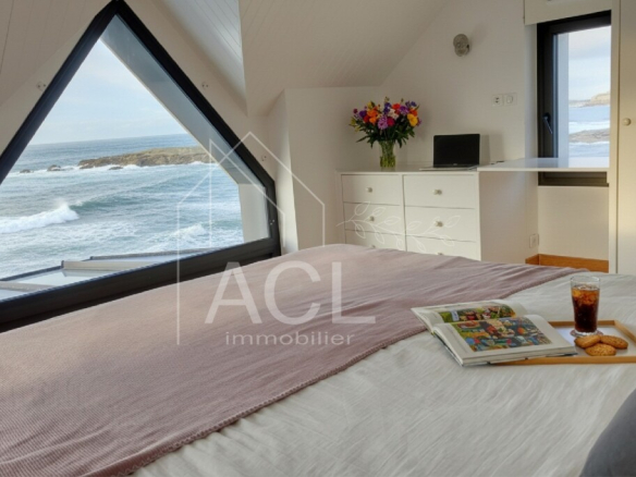 Sea-facing bedroom with a large triangular window, a bed with a pink throw, and a tray holding a magazine, iced coffee, and cookies.