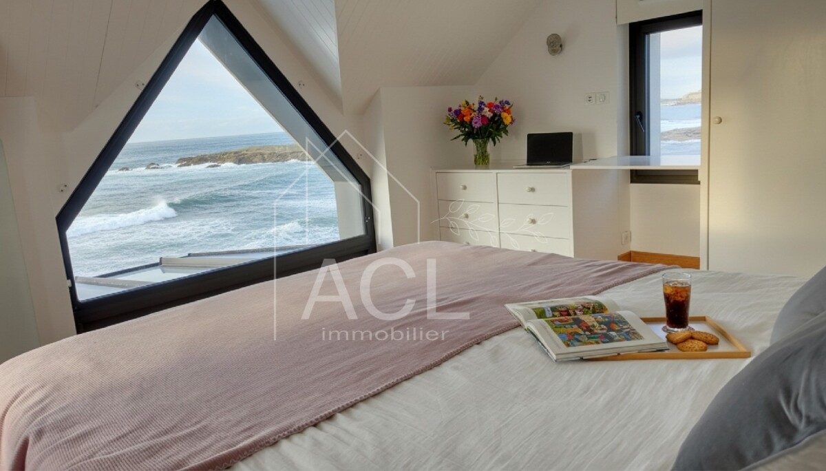 Sea-facing bedroom with a large triangular window, a bed with a pink throw, and a tray holding a magazine, iced coffee, and cookies.