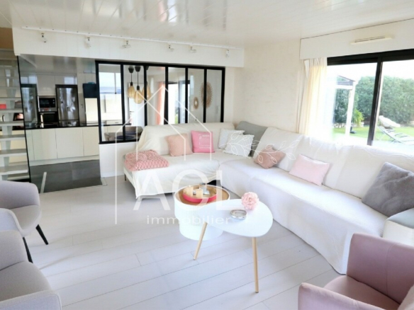 Bright open-plan living room with a white L-shaped sofa, pastel pink cushions, and a pale lavender accent chair; large glass doors to a green garden outside.